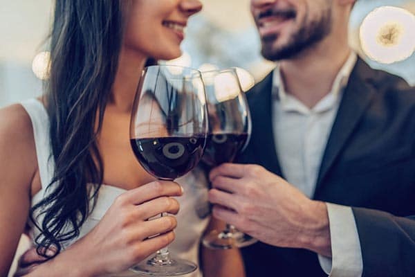 A couple dressed in formal attire clink glasses of red wine. They smile at each other, creating a warm and intimate atmosphere. The background is softly blurred with glowing lights, suggesting a romantic or celebratory setting.