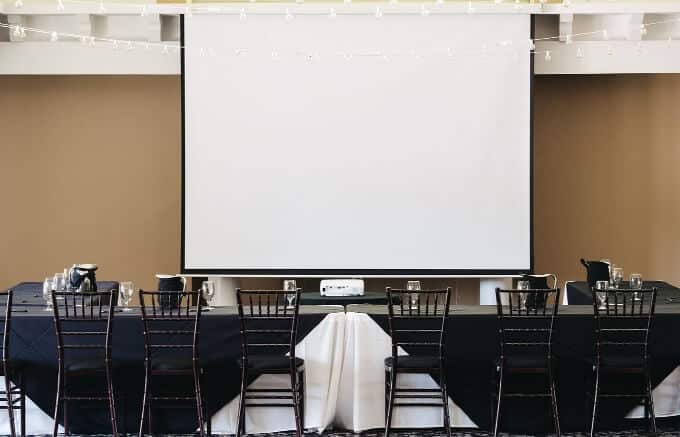 A large screen is displayed in a room furnished with chairs and tables, ready for a presentation or meeting.